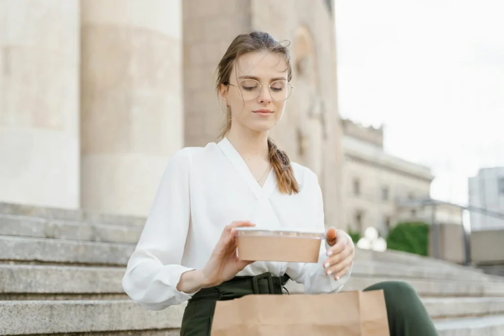 A young woman in a white blouse enjoys a takeaway lunch while sitting on urban steps.