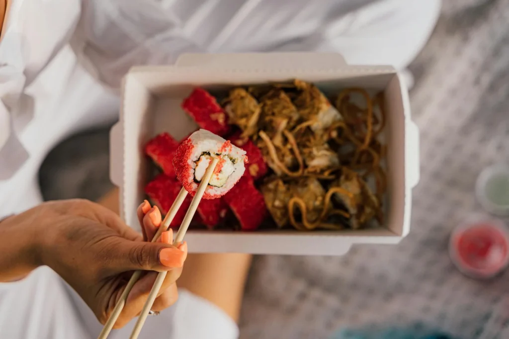 Close-up of sushi and noodles in a takeout box with chopsticks held by a hand.